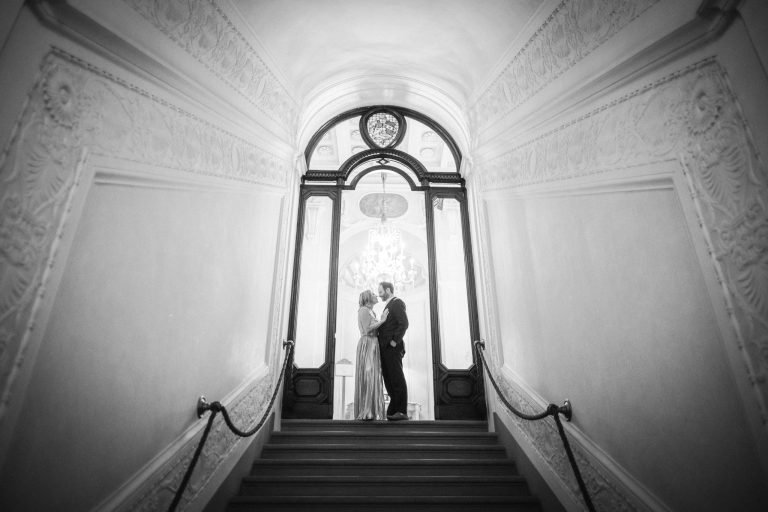 Black and white wide view of the couple kissing at the top of a symmetrical staircase